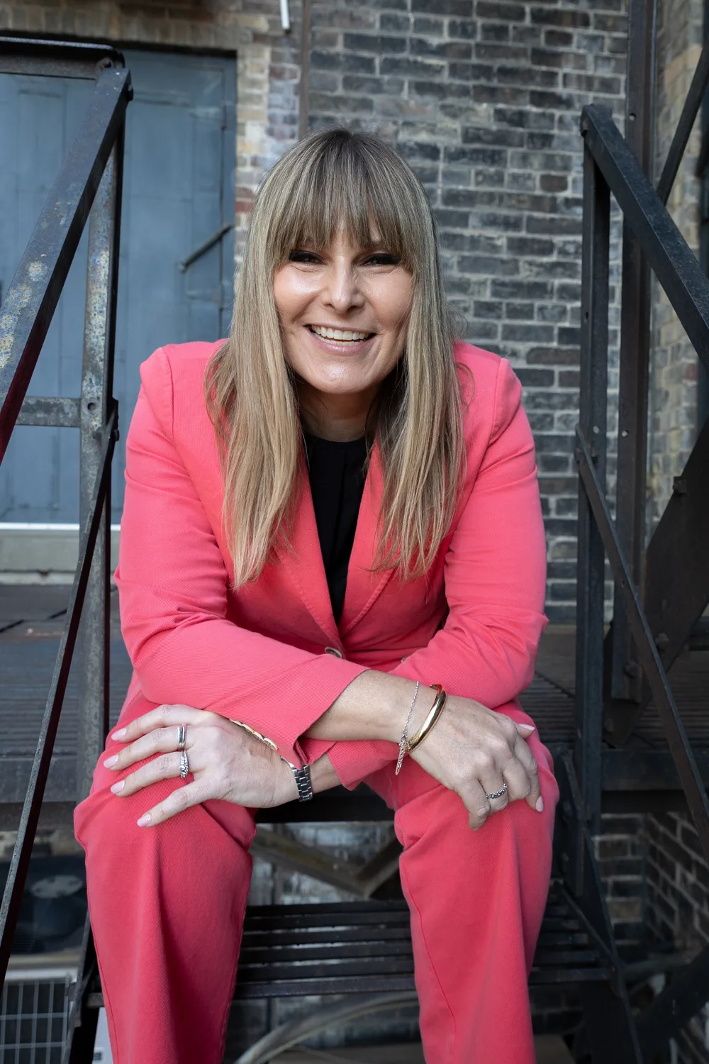 Maria sitting on steps in a coral suit, leaning forward with a warm smile